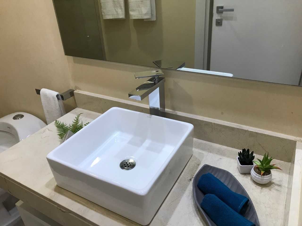 Une salle de bain moderne avec un grand lavabo blanc, un miroir, un porte-serviettes, un distributeur de savon et deux petites plantes en pot.
