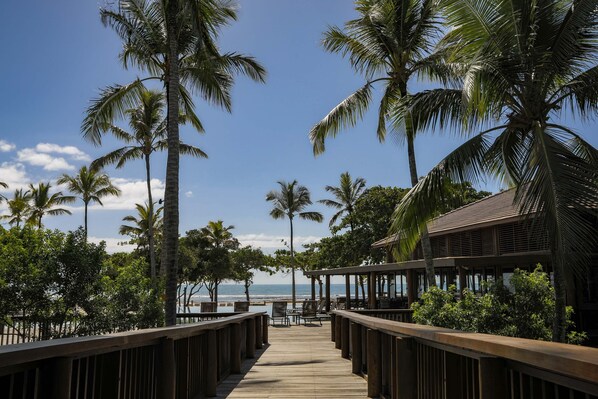 A wooden deck leading to a beachside area with palm trees and a building with a sloped roof. - Kûara Hotel (Porto Seguro)