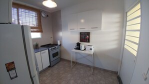 Cocina compacta con armarios blancos, estufa a gas, horno, una mesita con cafetera y un letrero en la pared.