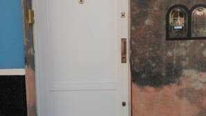 Una puerta blanca con un pequeño ventanal circular y un timbre. La puerta está en una pared con el número '900' encima.