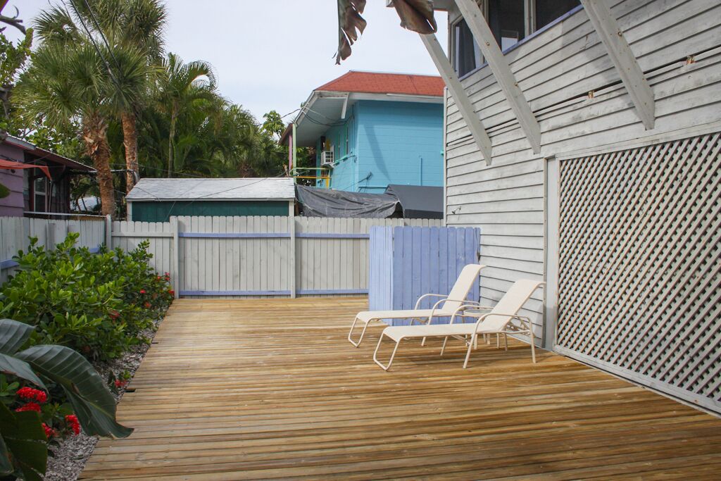 A wooden deck with two white lounge chairs, a blue house in the background, and a white fence.