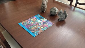 A dining table with a colorful puzzle mat, two ceramic animal figurines, and a wooden cabinet with framed pictures.
