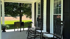 A covered porch with two rocking chairs and a small table, a potted plant, and a view of a baseball field.
