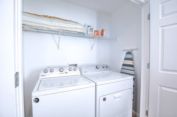 A laundry room with two stacked Whirlpool washers and dryers, a metal shelf above with folded clothes and laundry supplies. - North Shore- Family and Business Retreat (Lehi)