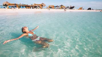 Eine Person treibt in klarem türkisblauem Wasser, im Hintergrund ein Strand und Hütten mit Reetdach.
