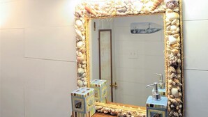 Une salle de bain avec un miroir encadré de coquillages, deux appliques murales et une étagère en bois où sont rangés les articles de toilette.
