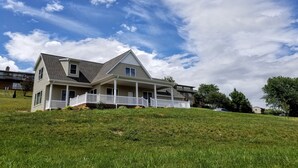 A two-story house with a front porch and a sloped roof, situated on a grassy hill with a clear sky.