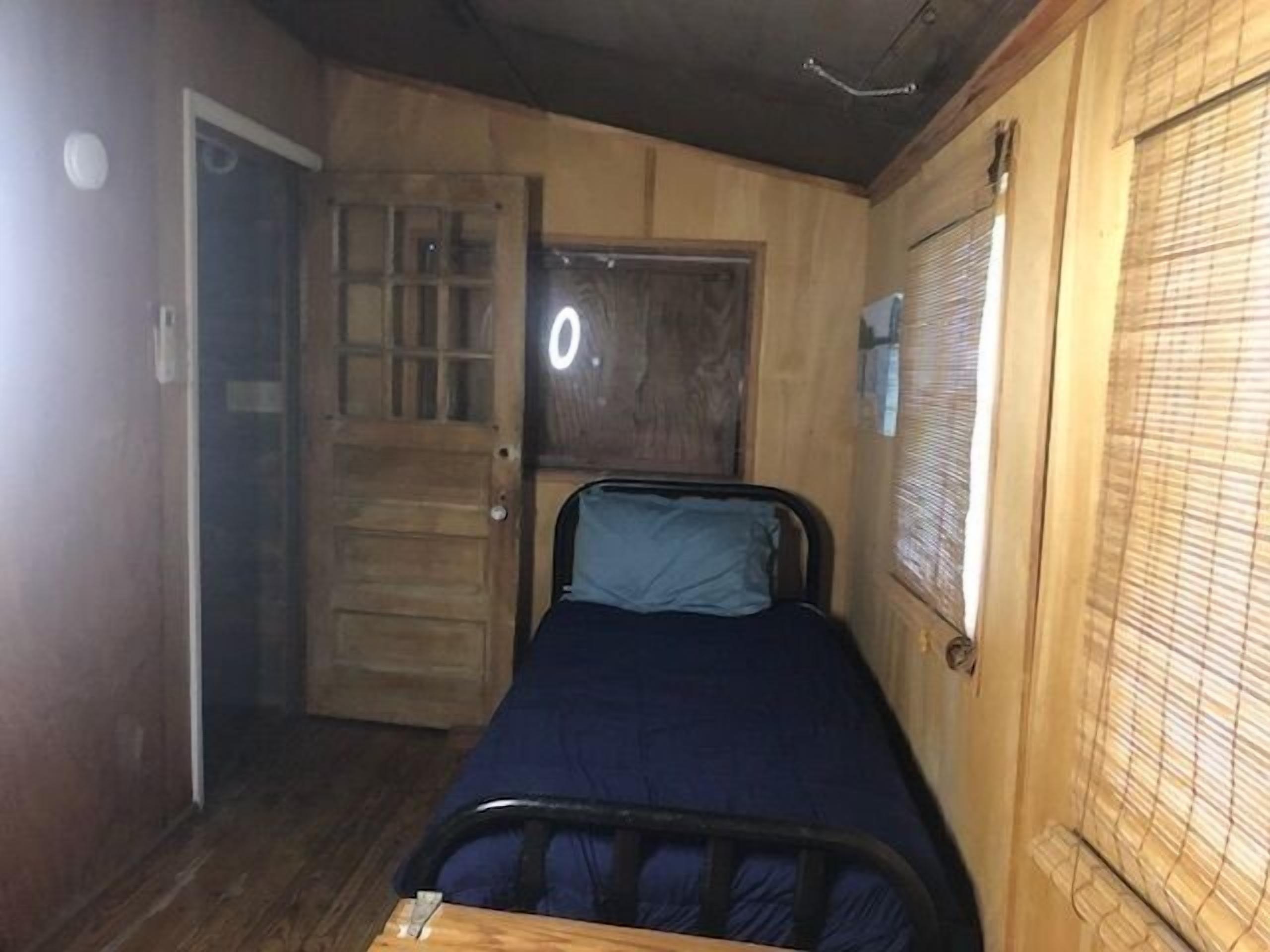 A small, single-bed room with a wooden door, a window with blinds, and a small table.