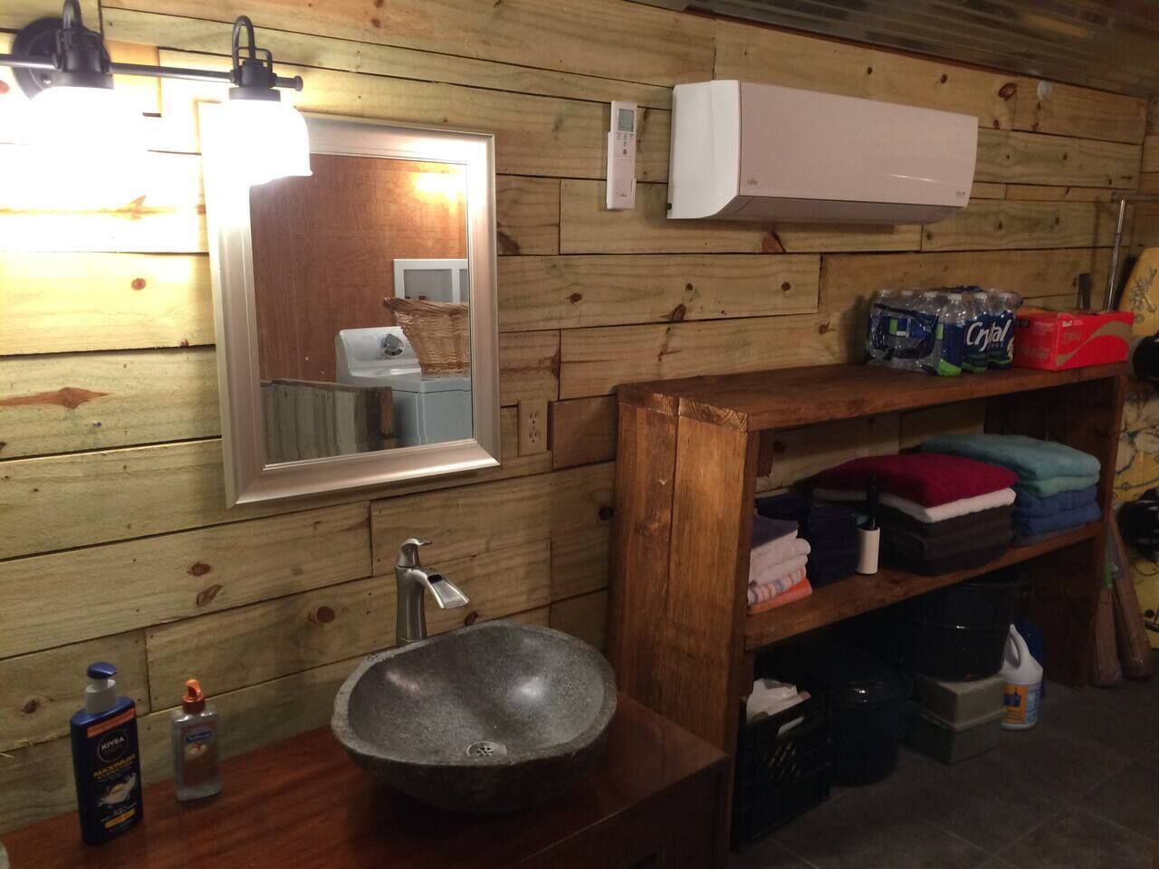 Baño de cabaña de madera con lavamanos de piedra, un espejo, un estante con artículos de tocador y un aire acondicionado blanco.