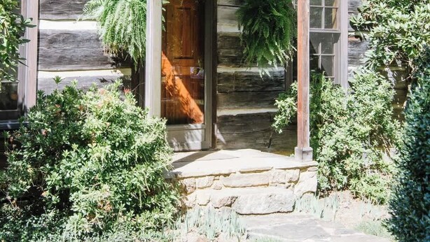 Een rustieke blokhut met een houten veranda, een stenen fundering en een schoorsteen.