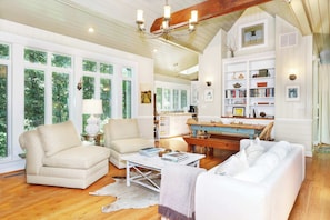 A spacious living room with large windows, a wooden ceiling, and a cozy seating area with a coffee table and books.