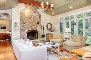 A cozy living room with a stone fireplace, checkered sofas, and a large window with a view of greenery.