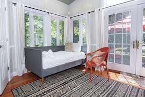 A bedroom with a gray sofa bed, a red chair, a patterned rug, and large windows with curtains.