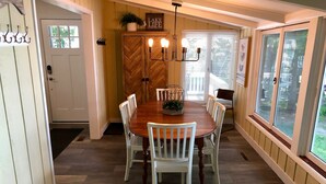 A dining area with a wooden table and white chairs, a wooden cabinet with a plant, and a window with blinds. - Brower Cottage - Lakefront Secluded Wooded Lot on Hutchins Lake Near Saugatuck (Fennville)
