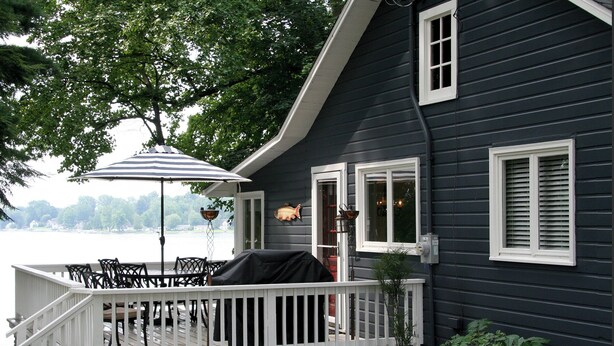 A dark blue house with a white deck, a grill, and a table with chairs. - Brower Cottage - Lakefront Secluded Wooded Lot on Hutchins Lake Near Saugatuck (Fennville)