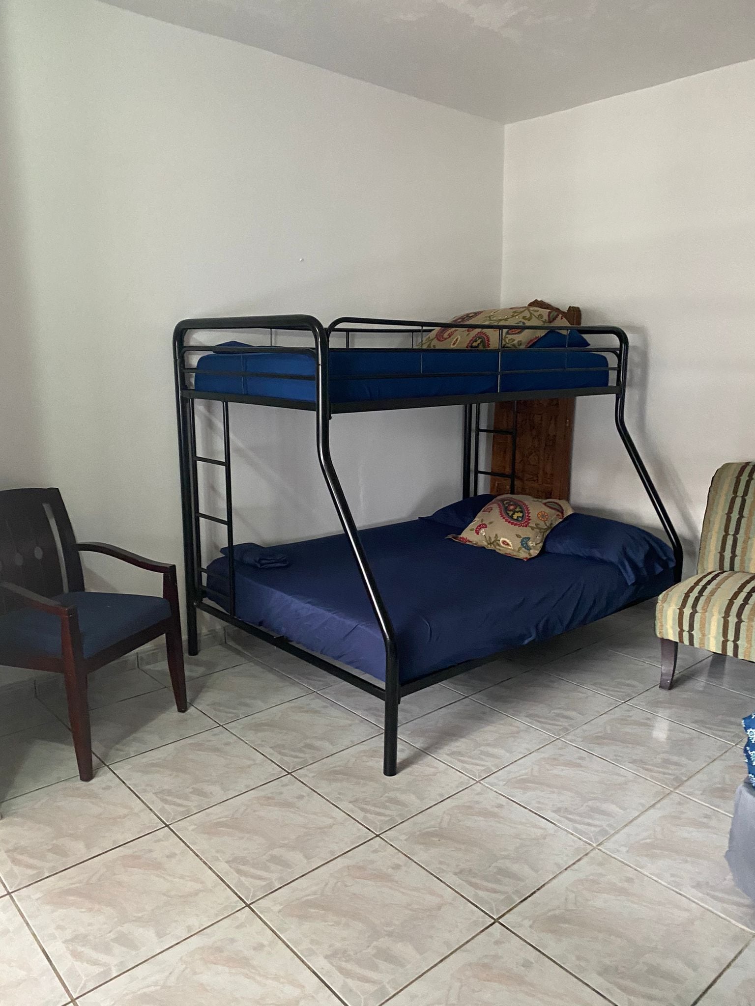 A bunk bed with a wooden headboard, a chair, and a striped armchair in a room with tiled flooring.