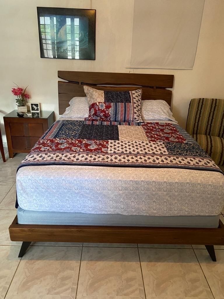 Un letto con copriletto decorato, una testiera in legno e un comodino con un vaso e una cornice.