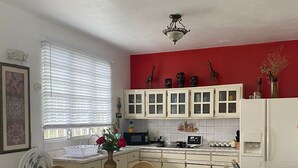 Une cuisine avec des meubles blancs, un mur d’accent rouge et un coin repas avec des chaises en osier et une table en verre.