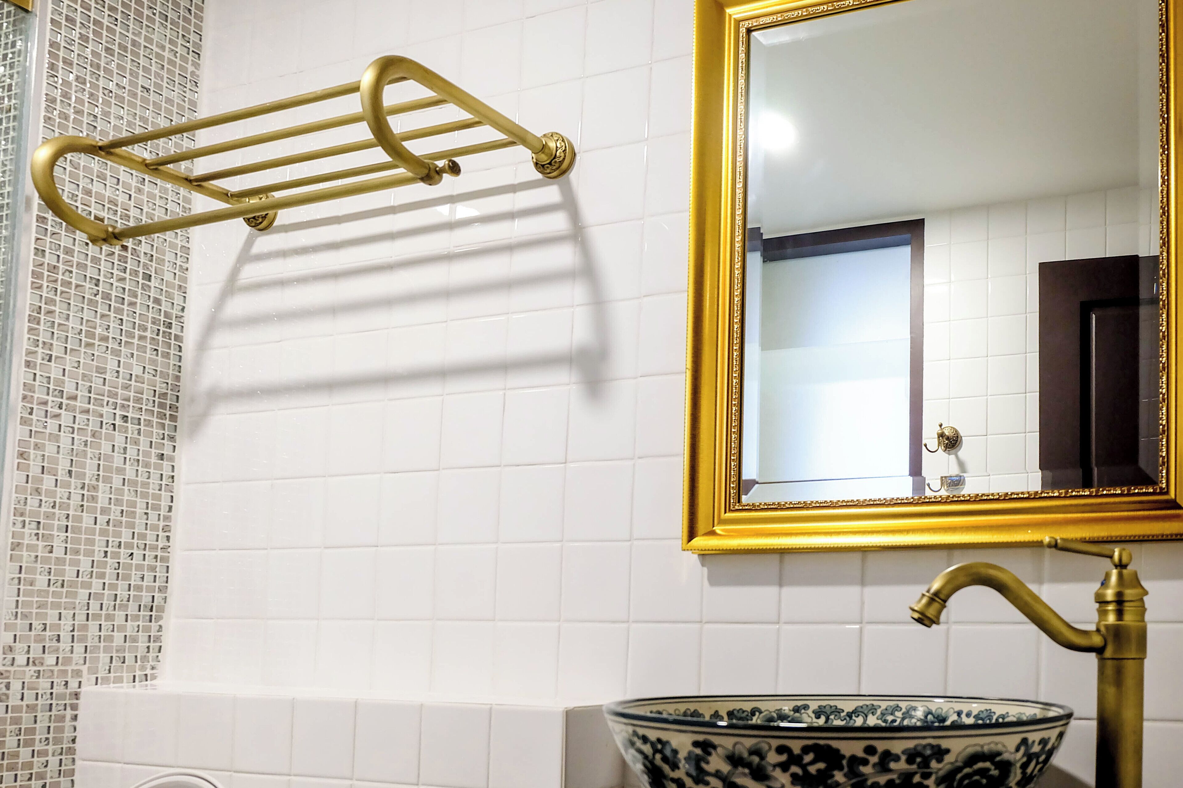 Une salle de bain avec des murs recouverts de carreaux blancs, un miroir à cadre doré, un porte-serviettes doré et un lavabo décoratif en forme de bol.
