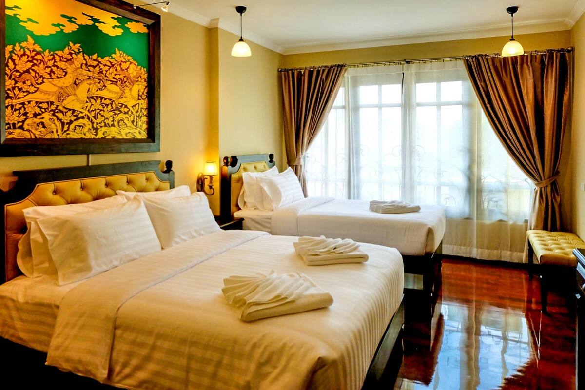 A hotel room with two beds, a large painting on the wall, and a view of the outdoors through the window.