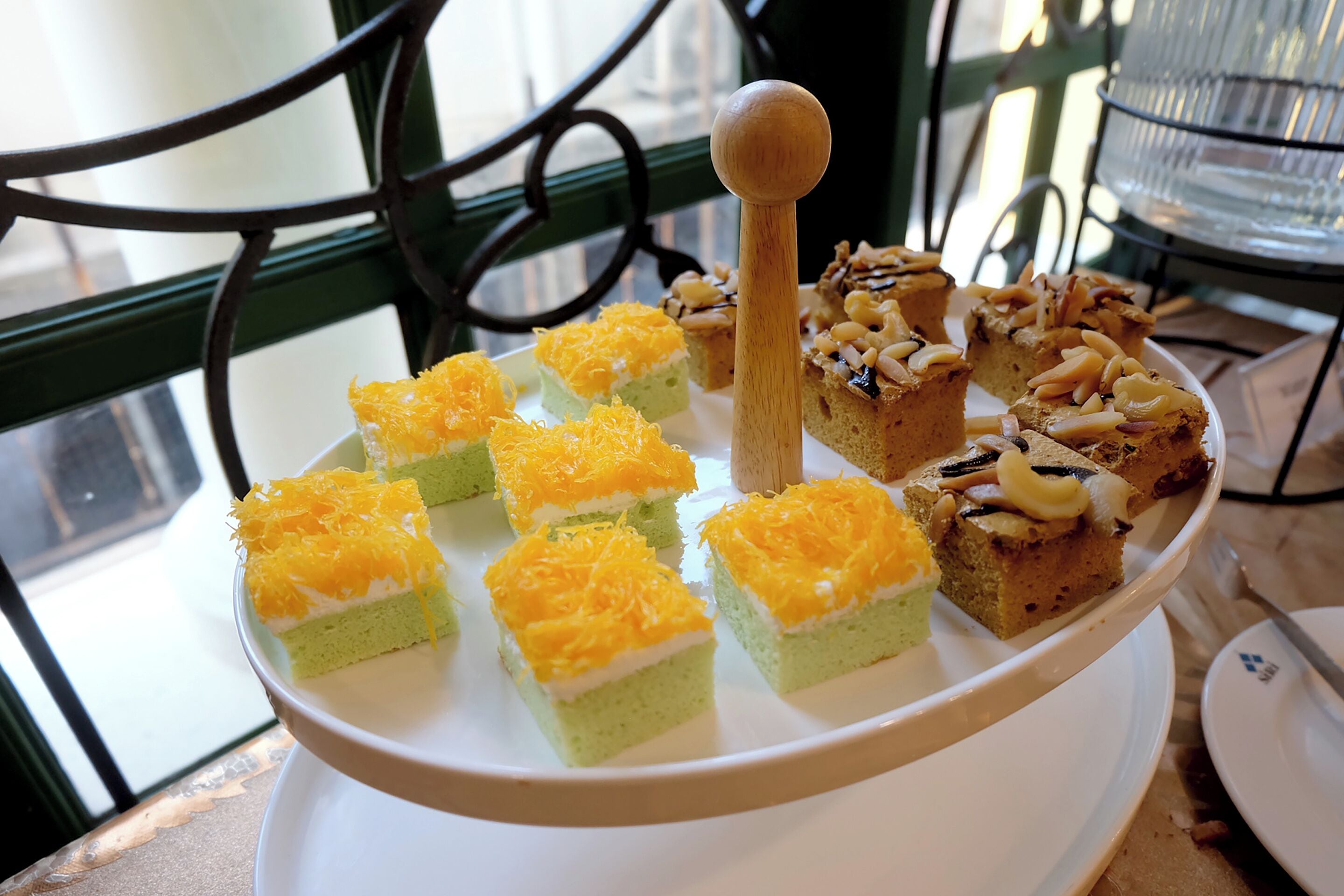 A tiered serving tray with various desserts, including cakes and pastries, on a table near a window with a metal railing.