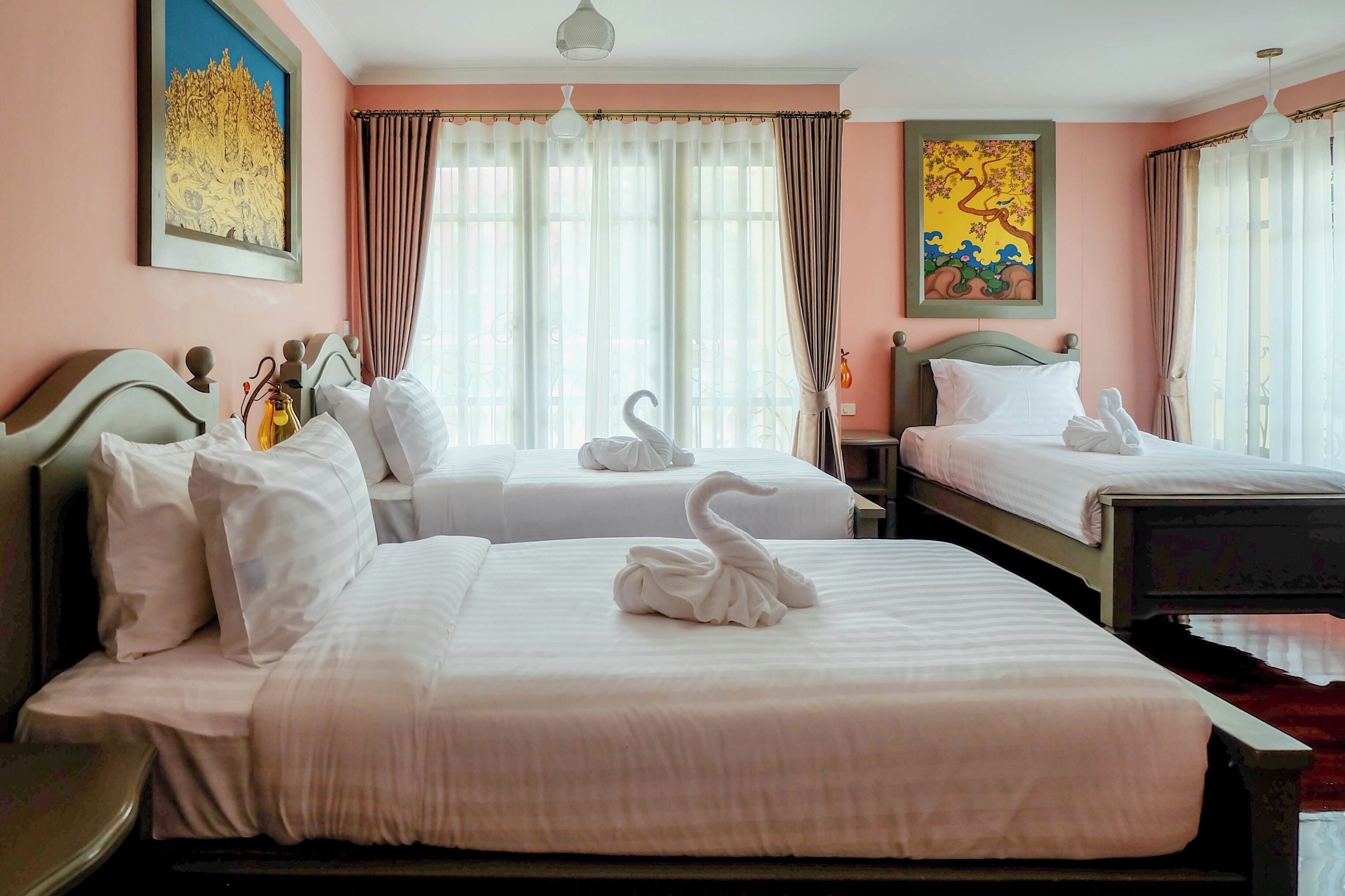Quarto de hotel com duas camas, cada uma com roupa de cama branca e almofadas de cisne decorativas. Há janelas amplas com cortinas leves, um quadro na parede e uma luminária no teto.
