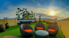 A rooftop terrace with wicker furniture and artificial grass, overlooking a cityscape with palm trees and a sunset. - Siri Heritage Bangkok Hotel (Bangkok)