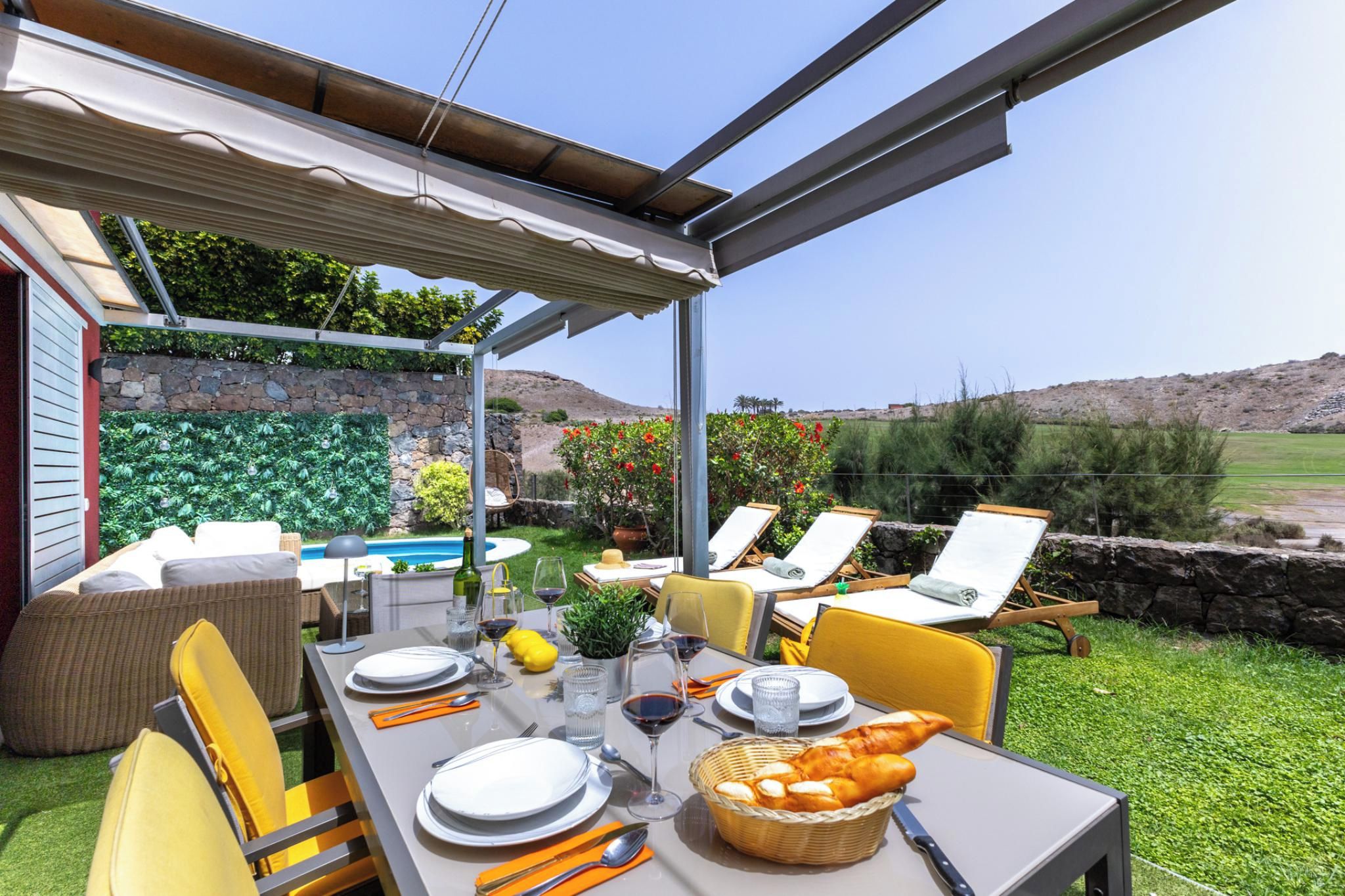 An outdoor dining area with a table set for two, surrounded by chairs, a pool, and a stone wall with greenery.