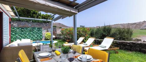 An outdoor dining area with a table set for two, surrounded by chairs, a pool, and a stone wall with greenery.