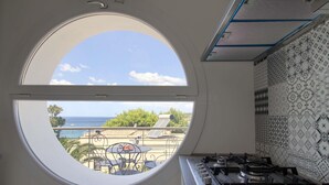 Cozinha com janela redonda que oferece vista para o oceano e um pátio com mobiliário de exterior.