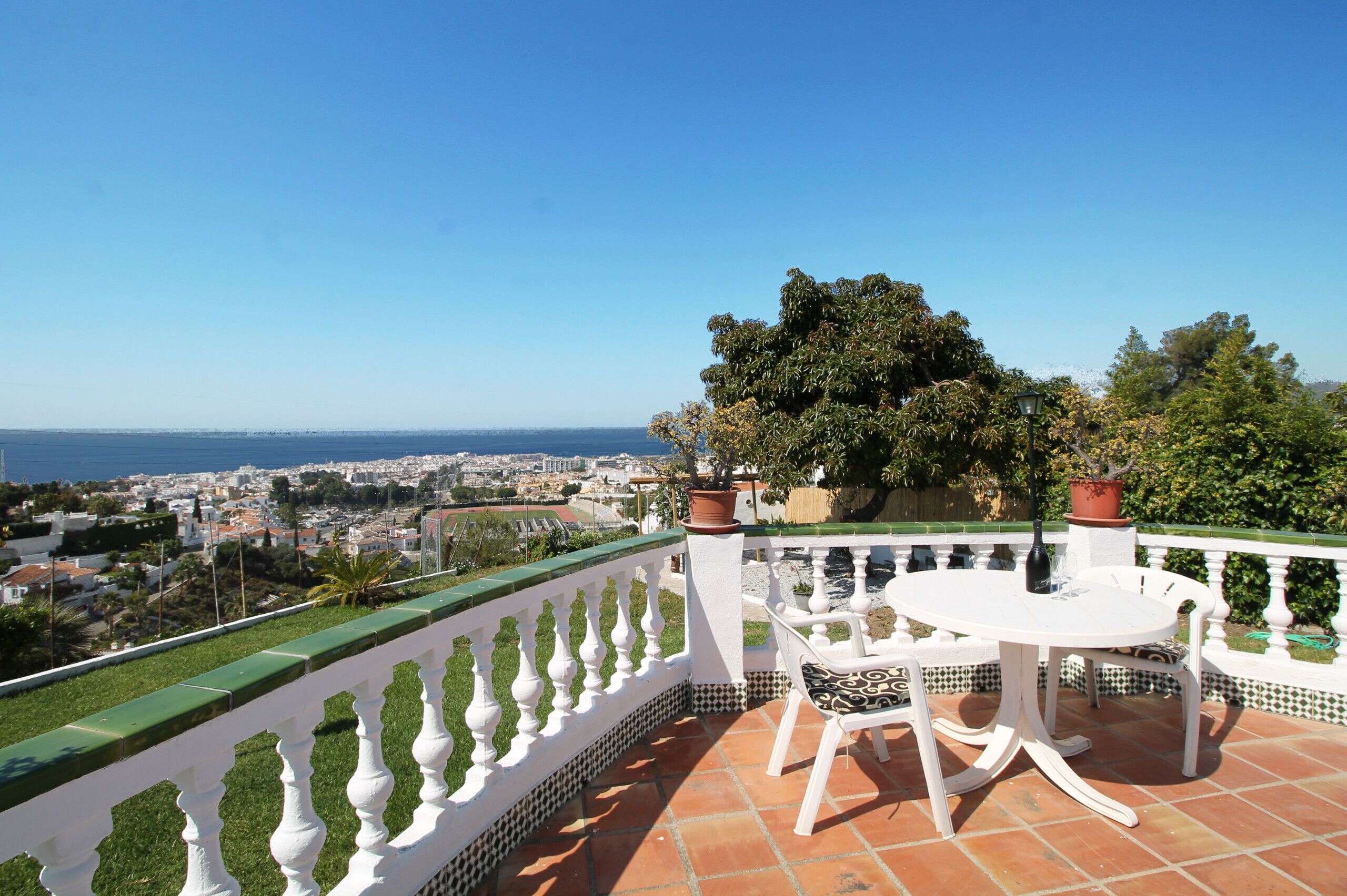 Een terras met een witte balustrade, een ronde tafel met stoelen en uitzicht op een kuststad.