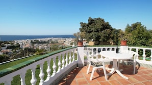 Eine Terrasse mit weißer Balustrade, einem runden Tisch mit Stühlen und Blick auf eine Küstenstadt.