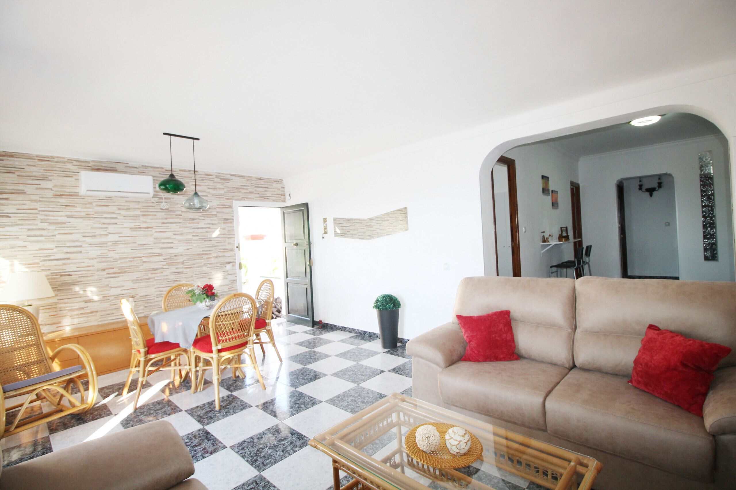 Una sala de estar con piso a cuadros, un sofá color beige con cojines rojos, sillas de mimbre, una mesa de centro de vidrio y una mesa de comedor con cojines rojos.