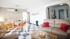 Ein Wohnzimmer mit Schachbrettmuster-Boden, einem beigen Sofa mit roten Kissen, Korbstühlen, einem Glastisch und einem Esstisch mit roten Kissen.