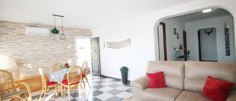 Una sala de estar con piso a cuadros, un sofá color beige con cojines rojos, sillas de mimbre, una mesa de centro de vidrio y una mesa de comedor con cojines rojos.