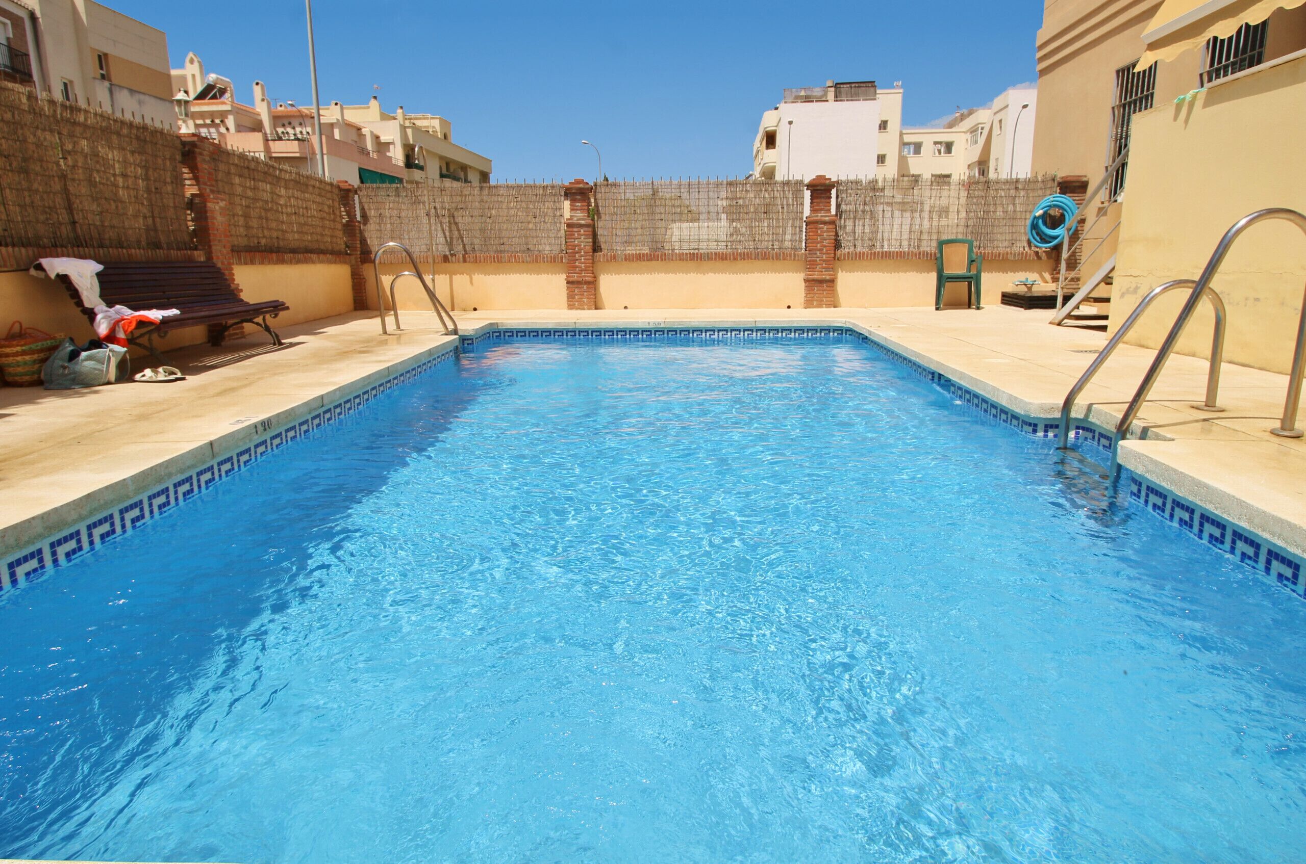 Una piscina rettangolare con superficie d'acqua cristallina azzurra, circondata da un basso muro in mattoni e una recinzione metallica.