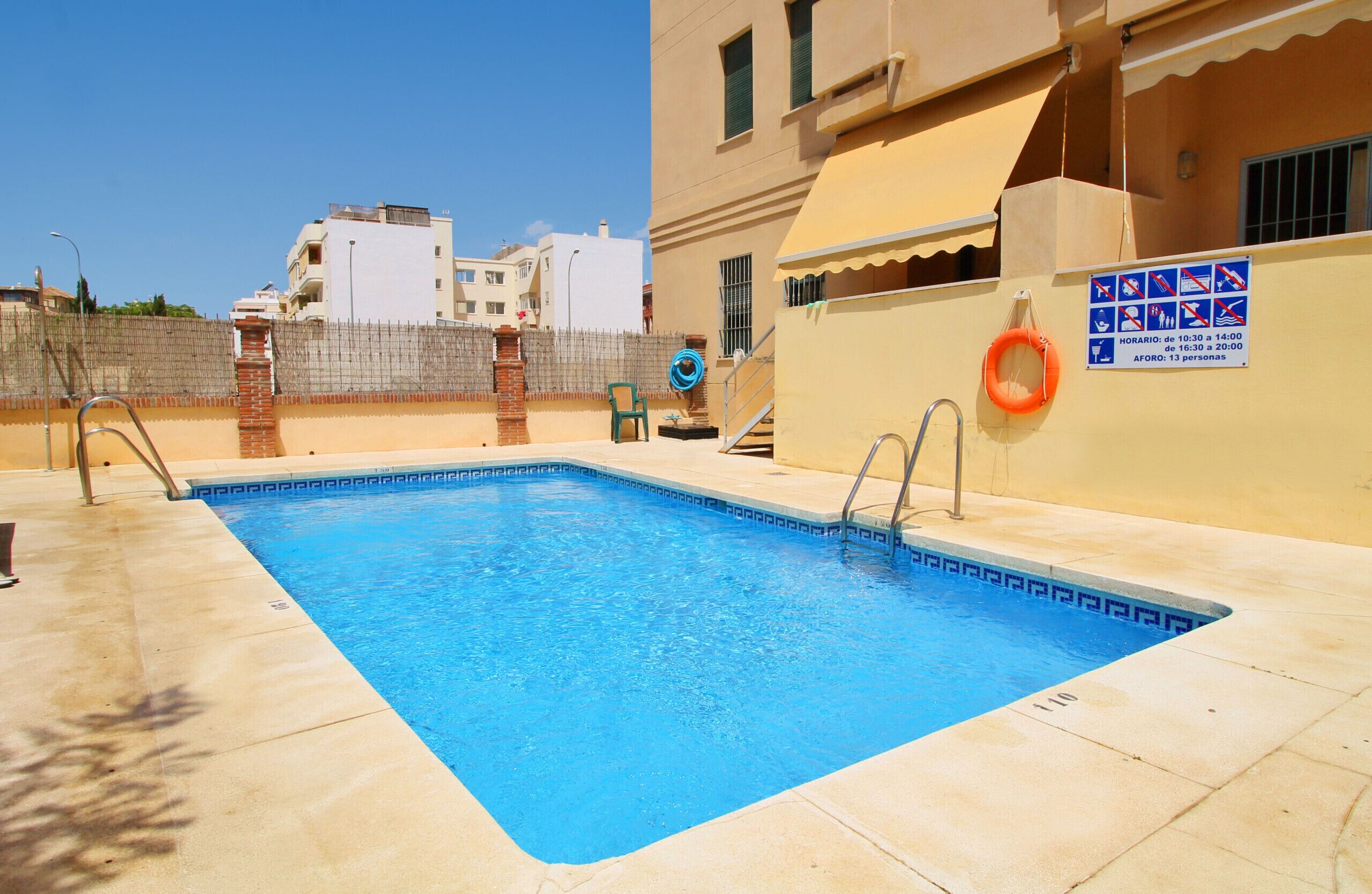 A rectangular swimming pool with a tiled edge, a lifebuoy attached to the wall, and a sign indicating a 13-person capacity.
