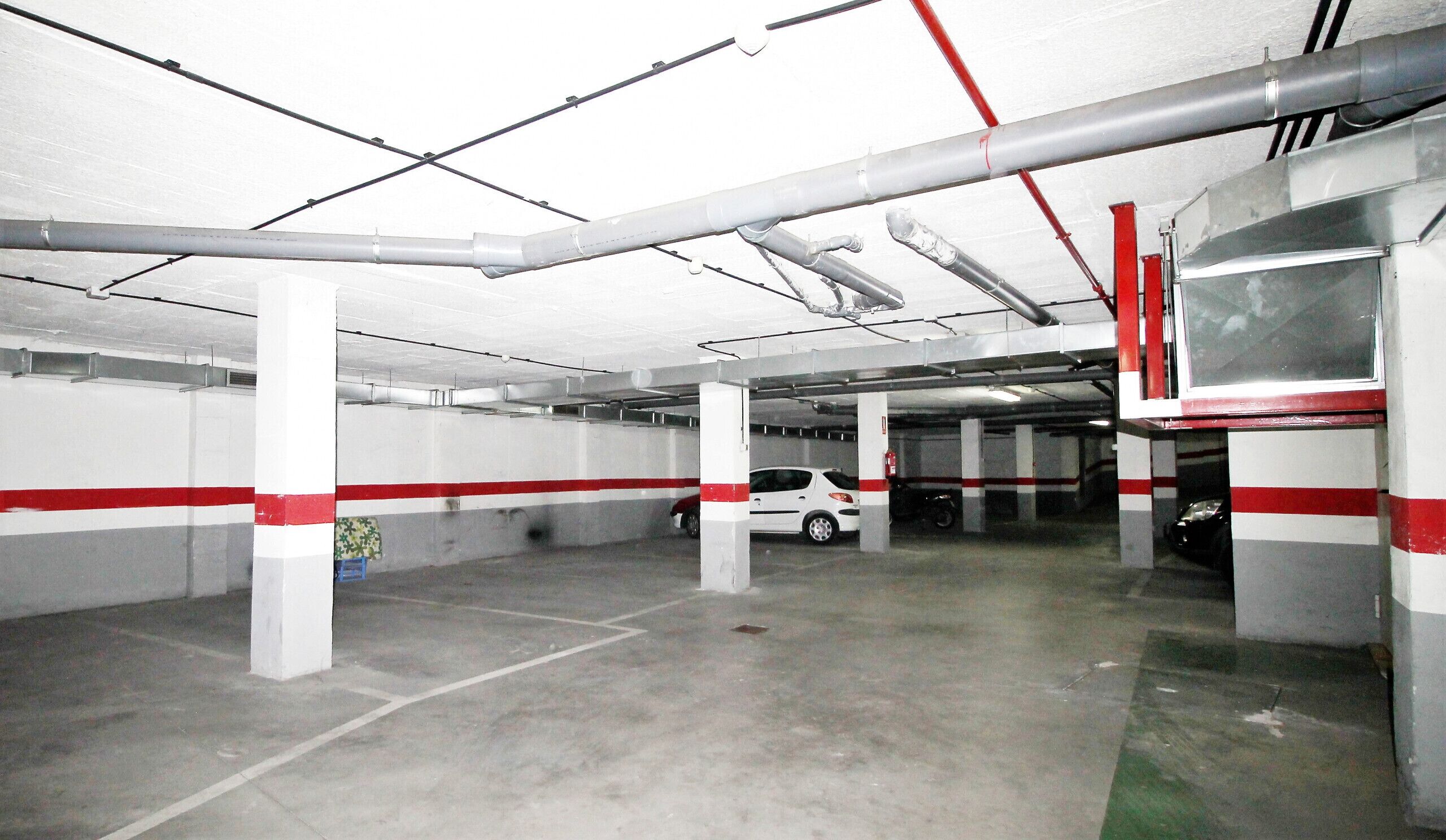 Um estacionamento multi andar com paredes listradas em branco e vermelho, pisos de concreto, e vigas e tubulações estruturais visíveis.