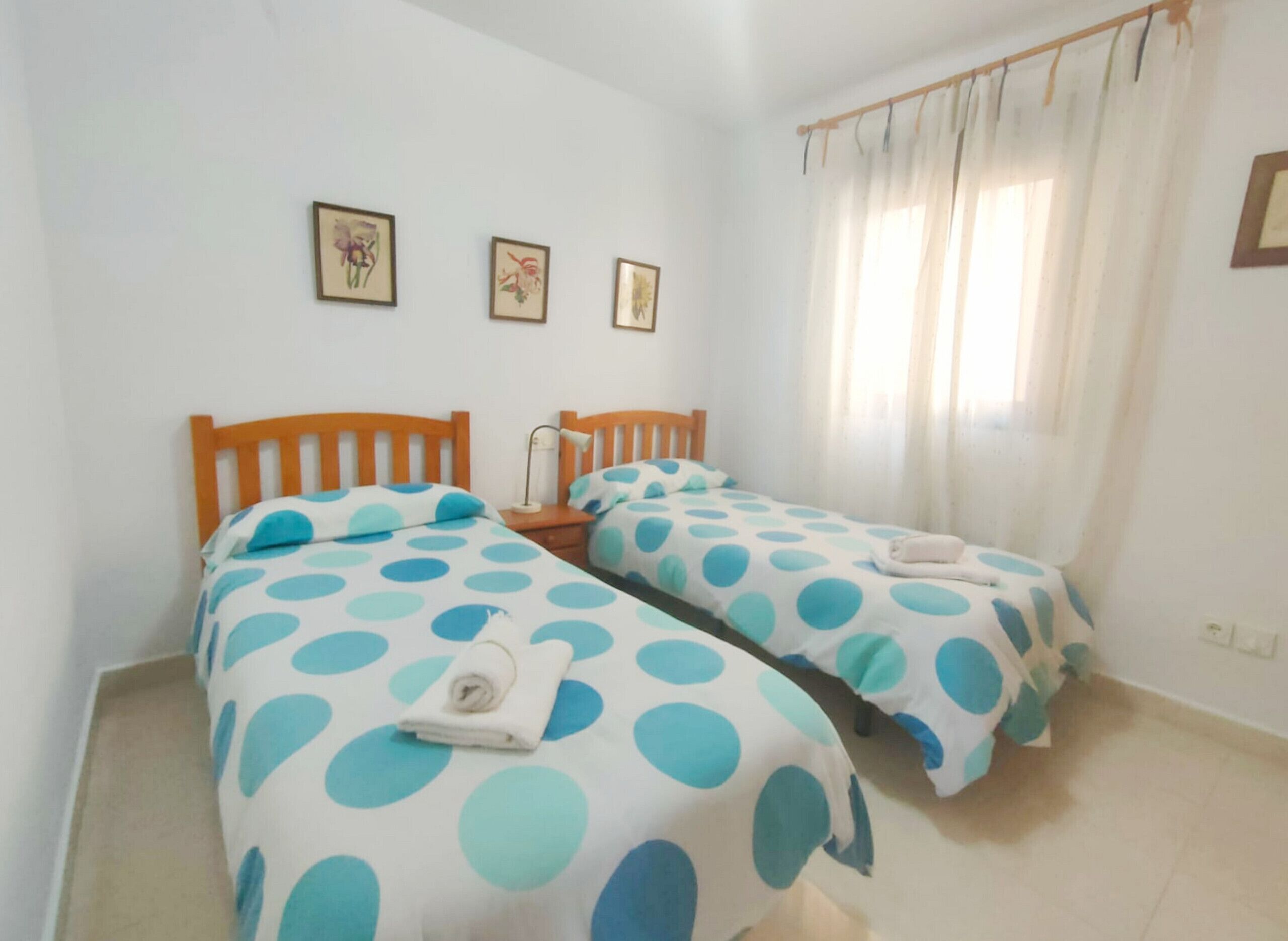 Ein Schlafzimmer mit zwei Betten, beide mit einer Betttdecke mit Punkten, einem Holz-Kopfteil und einem Nachttisch mit Lampe. An der Wand hängen drei gerahmte Bilder.