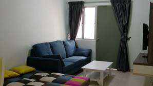 A living room with a blue sofa, a white coffee table, and a colorful ottoman. There is a ceiling fan and a window with curtains. - Goodlife Paradise Cameron Jaya (Tanah Rata)