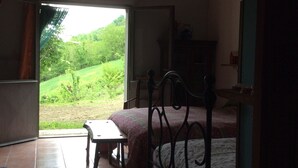 Un dormitorio con cama, una mesita y un banco. Se ve un paisaje verde a través de una puerta abierta.