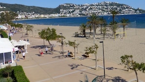 A beach with palm trees, a volleyball net, and a view of the sea and a hill with buildings. - Amazing apartment on the seafront in Roses Inmosantos Oasis D4 (Rosas / Roses)