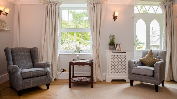Una habitación con dos sillones, una mesita con un florero y libros, y un ventanal con cortinas.