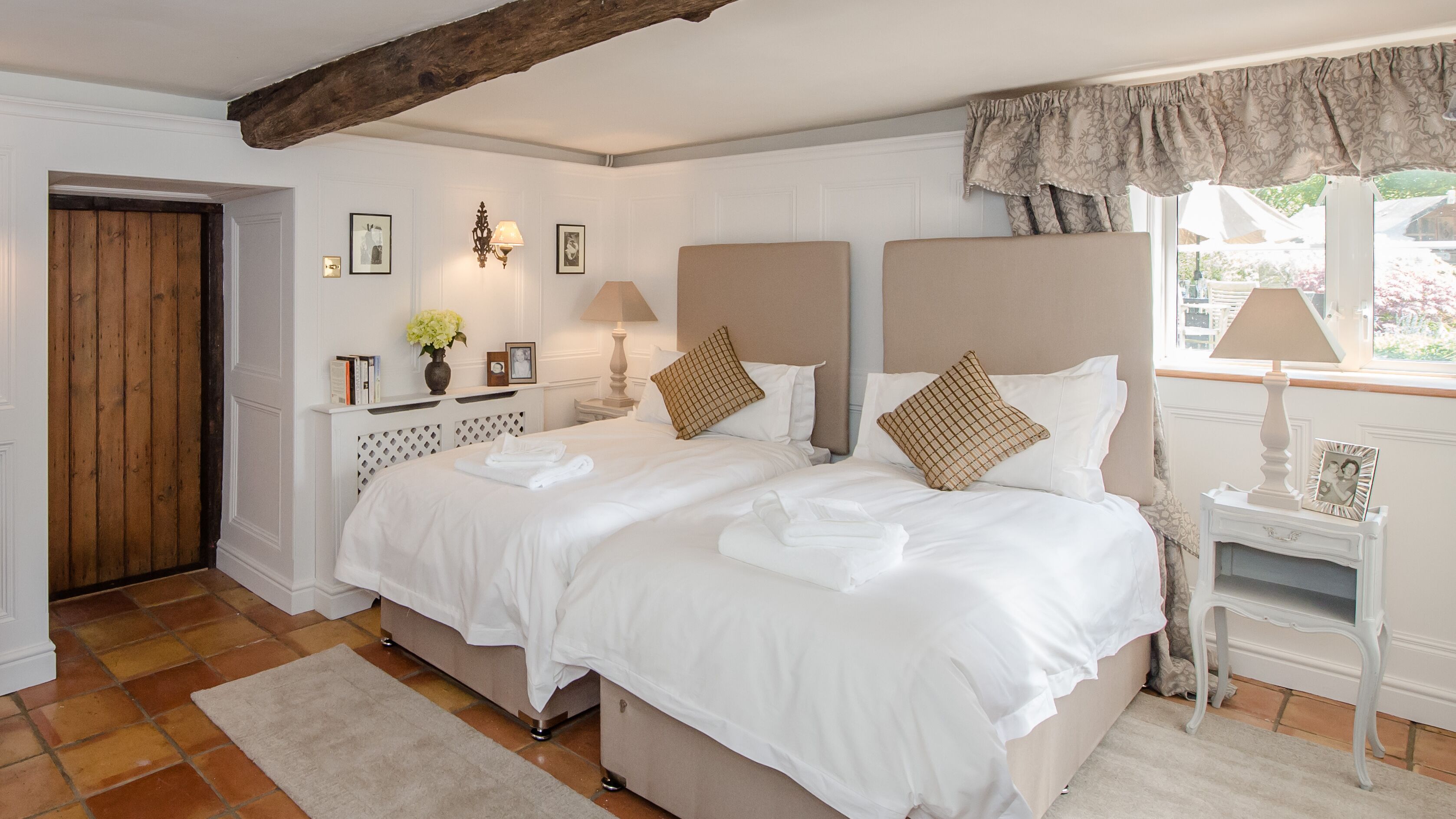 A neatly arranged bedroom with a large bed, bedside tables, a nightstand, a window with curtains, and a wooden door.