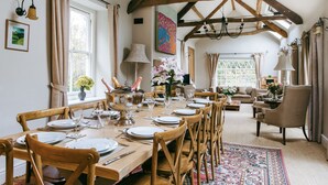 A dining area with a wooden table set for six, surrounded by wooden chairs. There is a sofa and a chair in the background, and a window with curtains.