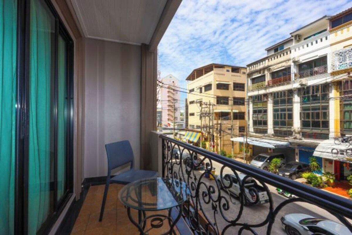 A balcony with a glass table and chair, overlooking a street with buildings and parked cars.
