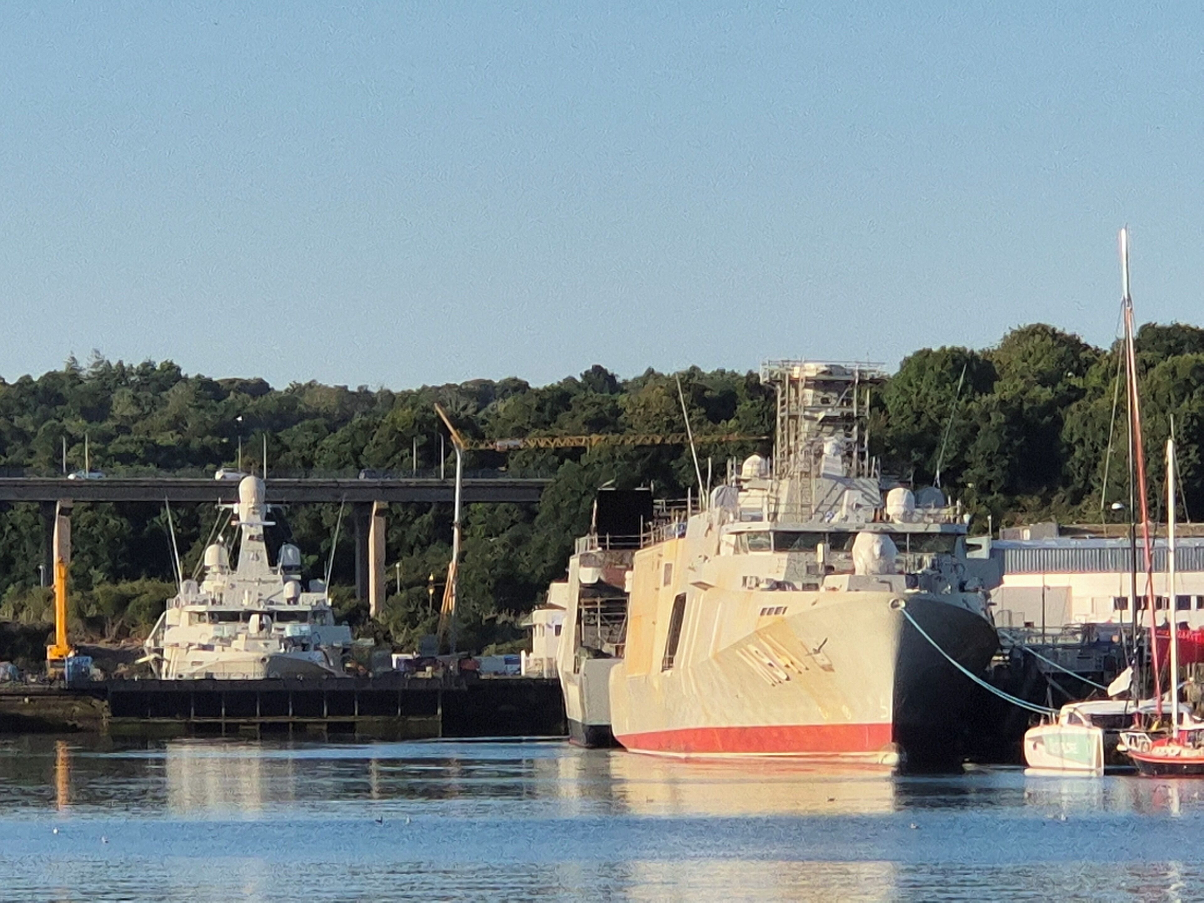 A large naval ship docked at a port with a bridge and greenery in the background.