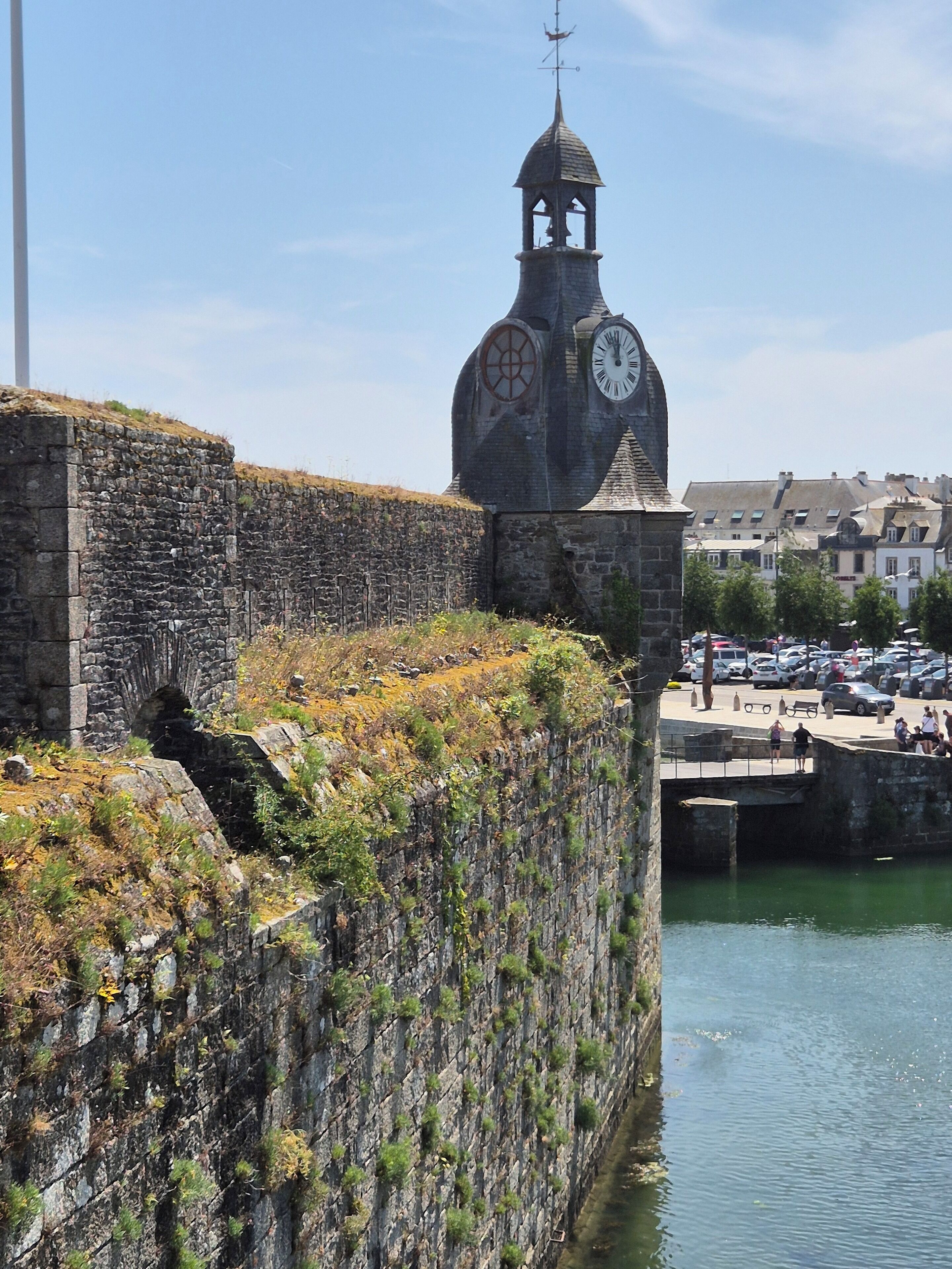 A historic stone wall with a clock tower, a bridge, and a body of water.