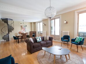 A spacious living room with a brown sofa, a round coffee table, and a chandelier. A spiral staircase is visible in the background. - Unique Penthouse Rooftop Terrace Puerta del Sol (Madrid)
