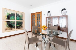 A dining area with a glass table, four chairs, a fruit bowl, and a wooden cabinet with glass doors.